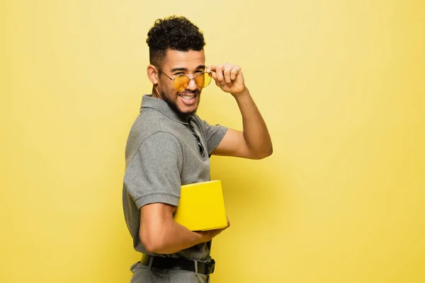 Young african american man adjusting sunglasses and holding book on yellow — Stock Photo