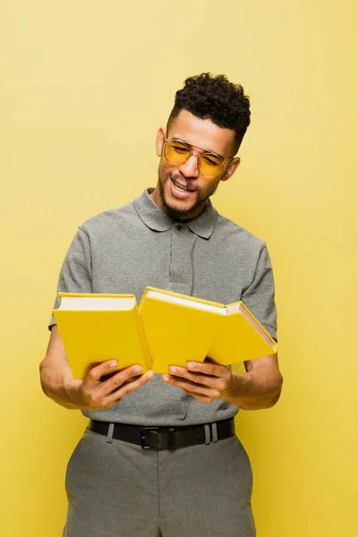Happy african american man in sunglasses and grey tennis shirt holding books isolated on yellow — Stock Photo