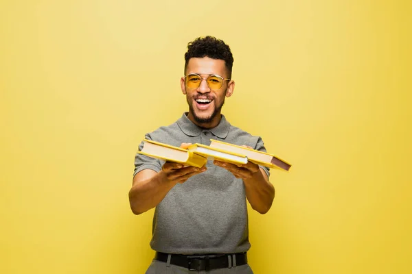 Alegre hombre afroamericano en gafas de sol y camisa de tenis gris sosteniendo libros en amarillo - foto de stock