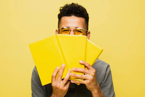 Homem americano africano em óculos de sol cobrindo rosto com livros isolados em amarelo — Fotografia de Stock