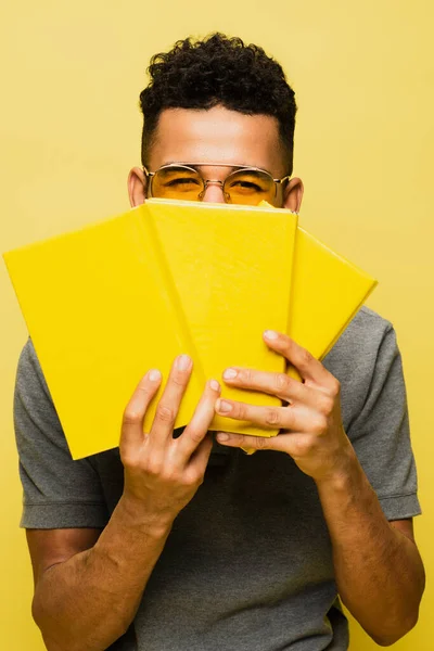 Homem americano africano em óculos de sol cobrindo rosto enquanto segurando livros isolados em amarelo — Fotografia de Stock