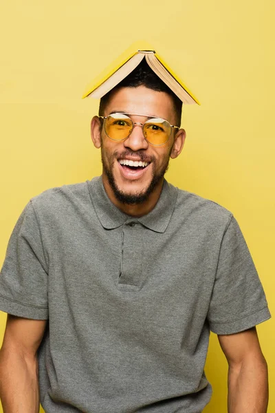 Hombre afroamericano feliz en gafas de sol y camisa de tenis gris con libro en la cabeza aislado en amarillo - foto de stock