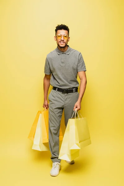 Piena lunghezza di felice uomo africano americano in occhiali da sole e camicia da tennis grigia che tiene borse della spesa su giallo — Foto stock