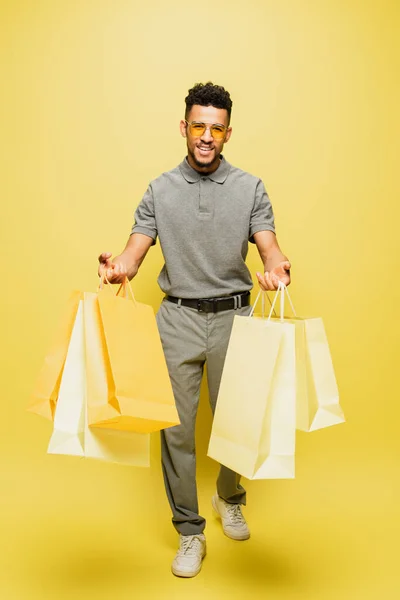 Longitud completa de hombre americano africano alegre en gafas de sol y camisa de tenis gris sosteniendo bolsas de compras en amarillo - foto de stock