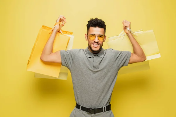 Щасливий афроамериканський чоловік в сонцезахисних окулярах і сіра тенісна сорочка тримає сумки на жовтому — стокове фото