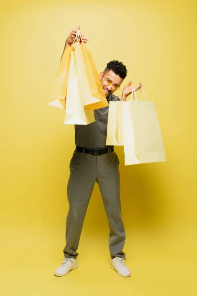 Full length of positive african american man in sunglasses and grey tennis shirt holding shopping bags on yellow — Stock Photo