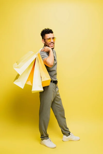 Homme afro-américain joyeux en lunettes de soleil et chemise de tennis grise tenant des sacs à provisions sur jaune — Photo de stock