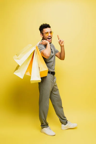 Piena lunghezza di felice uomo afroamericano in occhiali da sole e camicia da tennis grigia che tiene borse della spesa e punta sul giallo — Foto stock