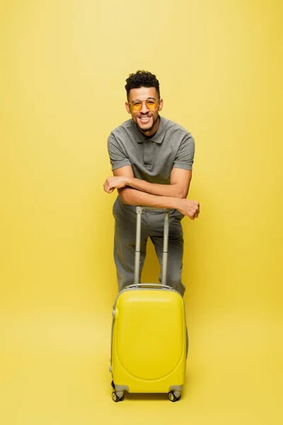 Comprimento total de homem americano africano feliz em óculos de sol e camisa de tênis cinza com bagagem em amarelo — Fotografia de Stock