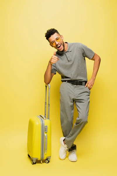 Homme afro-américain heureux en lunettes de soleil et chemise de tennis grise montrant pouce vers le haut près des bagages sur jaune — Photo de stock