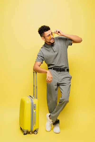 Toute la longueur de l'homme afro-américain élégant en lunettes de soleil et chemise de tennis grise debout près des bagages sur jaune — Photo de stock