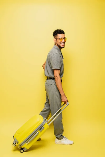 In voller Länge glücklicher afrikanisch-amerikanischer Mann mit Sonnenbrille und grauem Tennishemd, der mit Gepäck auf gelb läuft — Stockfoto