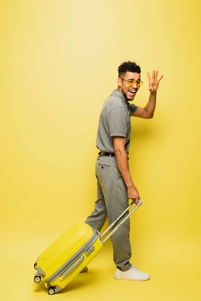 Longitud completa de hombre afroamericano feliz en gafas de sol y camisa de tenis gris caminando con el equipaje y agitando la mano en amarillo - foto de stock