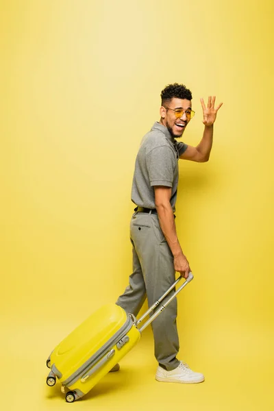 Full length of happy african american man in sunglasses and grey tennis shirt walking with luggage and waving hand on yellow — Stock Photo