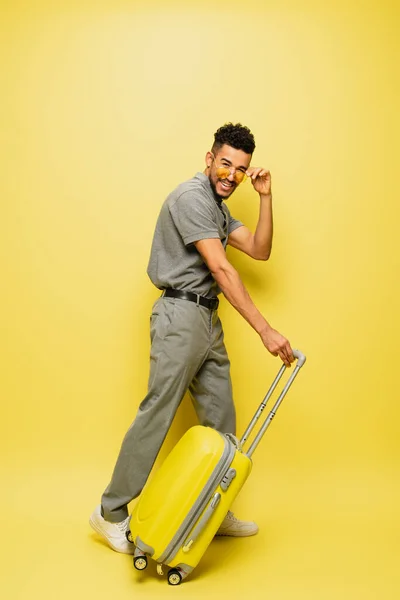 Full length of cheerful young african american man adjusting sunglasses and standing with luggage on yellow — Stock Photo