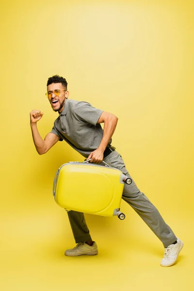 Longitud completa del hombre afroamericano excitado en gafas de sol y camisa de tenis gris llevando equipaje en amarillo - foto de stock