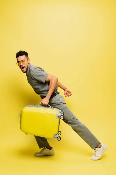 Comprimento total de homem americano africano espantado em óculos de sol e camisa de tênis cinza com bagagem em amarelo — Fotografia de Stock