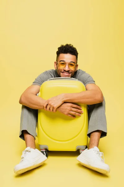 Longitud completa de hombre afroamericano feliz en gafas de sol sentado con equipaje en amarillo - foto de stock