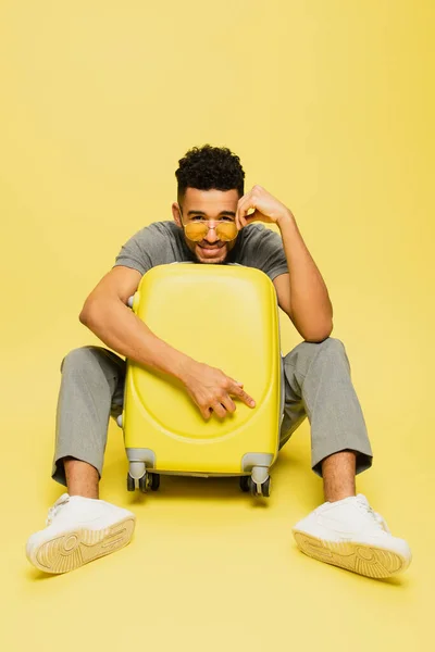 Pleine longueur d'homme afro-américain positif dans des lunettes de soleil assis avec des bagages sur jaune — Photo de stock