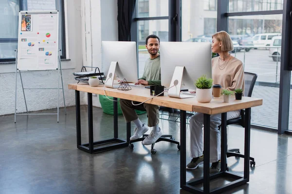 Complacidos colegas interracial buscando ta uno al otro mientras trabajaba en la oficina moderna - foto de stock