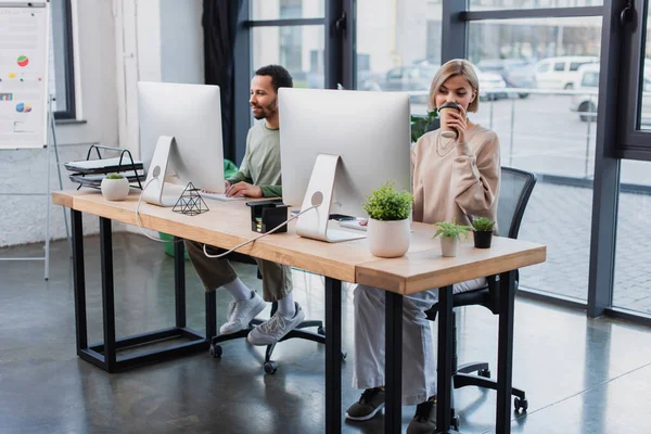 Blonde Frau trinkt Coffee to go und schaut auf Computerbildschirm in der Nähe eines afrikanischen amerikanischen Kollegen — Stockfoto