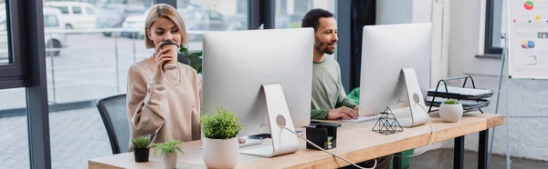 Rubia bebiendo café para ir y mirando el monitor de la computadora cerca de un colega afroamericano, pancarta - foto de stock