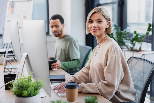 Blonde woman looking at camera near computer monitor and blurred african american colleague — Stock Photo