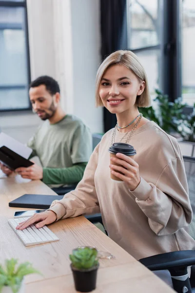 Cheerful blonde woman holding paper cup near blurred african american colleague — Stock Photo