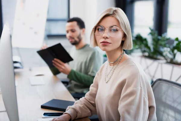 Blonde woman in glasses looking at camera near blurred african american colleague — Stock Photo