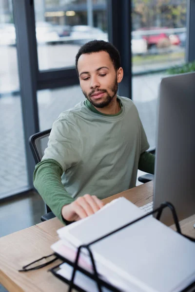 Barbudo afroamericano hombre mirando documentos en bandeja de documentos cerca borrosa monitor de la computadora - foto de stock