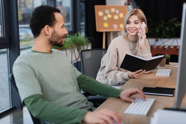 Mujer rubia sonriendo mientras habla en el teléfono inteligente y mirando a un colega afroamericano barbudo - foto de stock