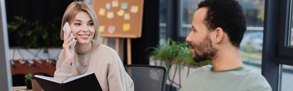 Mujer rubia sonriendo mientras habla en el teléfono inteligente y mirando a un colega afroamericano barbudo, pancarta - foto de stock