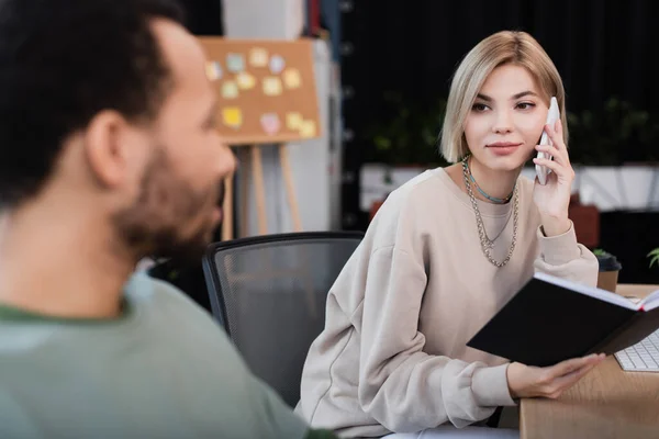 Rubia mujer hablando en el teléfono inteligente y mirando borrosa africana americana colega - foto de stock