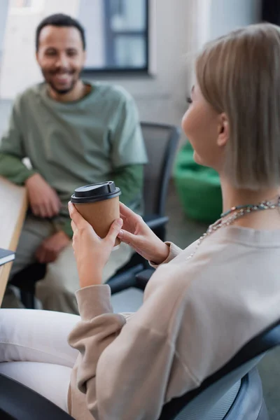 Rubia mujer sosteniendo papel taza cerca borrosa africano americano manager - foto de stock