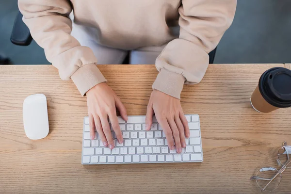 Vista dall'alto della donna d'affari digitando sulla tastiera del computer vicino tazza di carta e occhiali — Foto stock