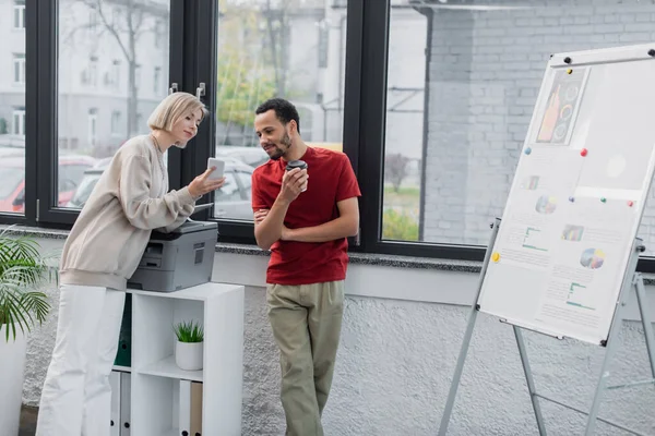 Blonde woman showing smartphone to african american colleague with paper cup near printer — Stock Photo