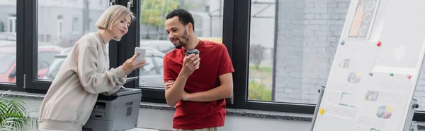 Blonde woman showing smartphone to african american colleague with paper cup near printer, banner — Stock Photo