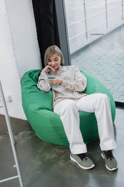 Gerente feliz sentado na cadeira saco de feijão e usando smartphone — Fotografia de Stock
