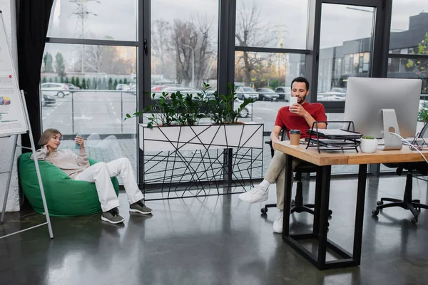 Femme blonde assise dans une chaise de sac de haricots et utilisant un smartphone près d'un collègue afro-américain au bureau — Photo de stock