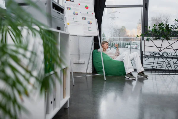 Blonde manager sitting in bean bag chair and using smartphone — Stock Photo