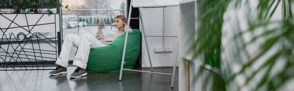 Blonde manager sitting in bean bag chair and using smartphone, banner — Stock Photo