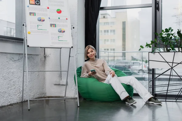 Smiling manager sitting in bean bag chair and holding smartphone — Stock Photo