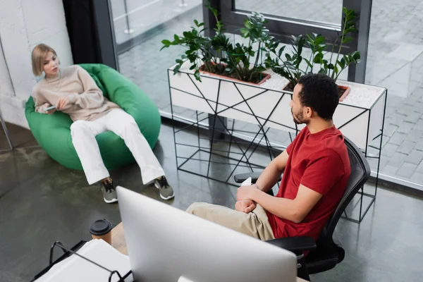 Vista de ángulo alto del gerente rubio sentado en la silla de la bolsa de frijol y sosteniendo el teléfono inteligente mientras habla con un colega afroamericano — Stock Photo