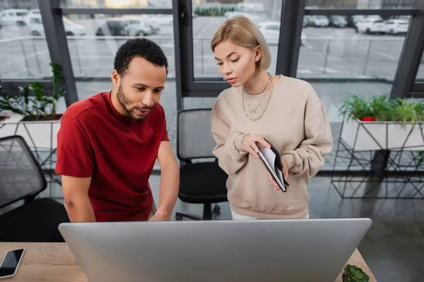 High angle view of blonde manager holding notebook near computer monitor and african american colleague — Stock Photo