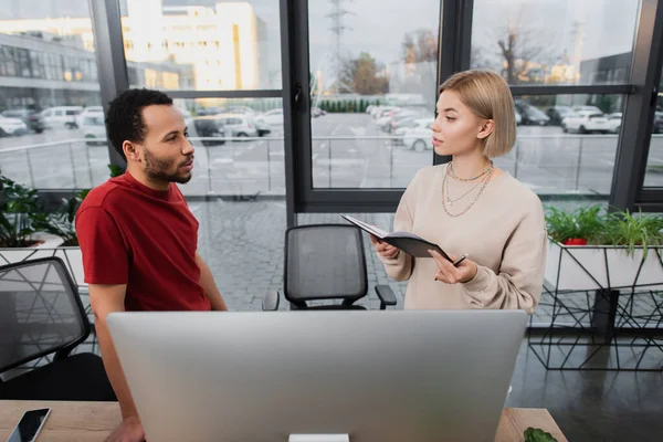 Rubia mujer de negocios sosteniendo portátil cerca del monitor de la computadora y colega afroamericano - foto de stock