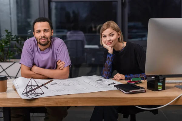 Young multiethnic interior designers smiling near colorful samples and blueprint — Stock Photo