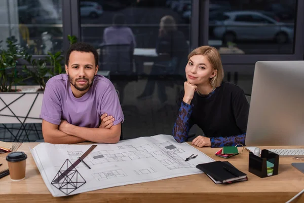 Junge interrassische Innenarchitekten in der Nähe bunter Muster und Baupläne auf dem Schreibtisch — Stockfoto