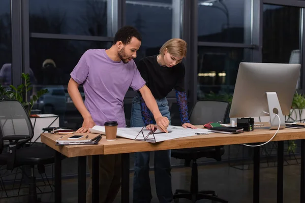 Young interracial colleagues looking at blueprint on desk — Stock Photo