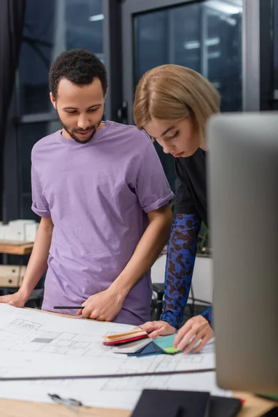 Multiethnic interior designers looking at colorful samples near blueprint — Stock Photo