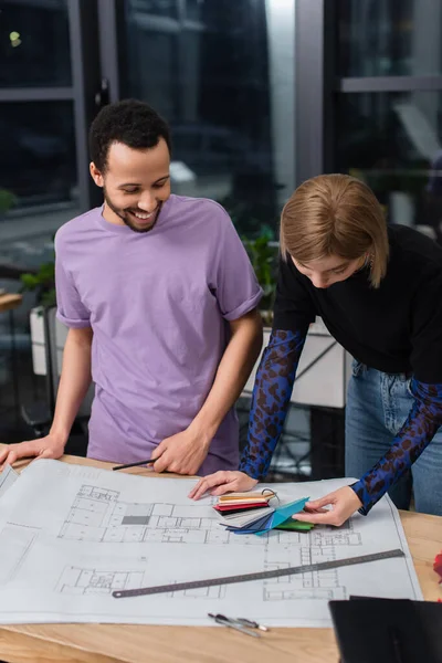 Alegre designer de interiores afro-americano olhando para amostras coloridas perto colega loira e blueprint na mesa — Fotografia de Stock